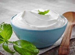 Creamy white natural yogurt in a blue bowl. A wooden spoon to the right and herbal leaves to the left.
