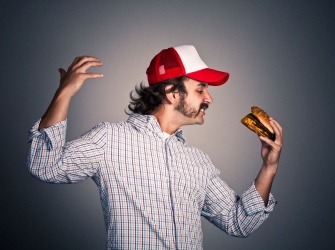 A man with a red and white basecap is holding a hamburger (Meat? Veggy? vegan?) in an artisitc way. He is looking forward to biting into it.